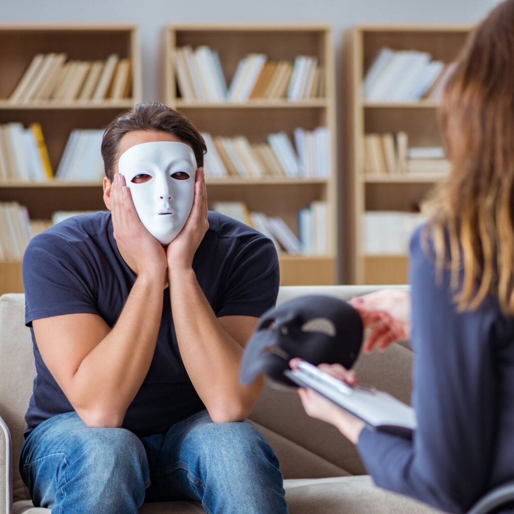 A distressed man sits on a couch, covering his face, while a woman offers him a mask and holds a clipboard. Bookshelves are in the background.