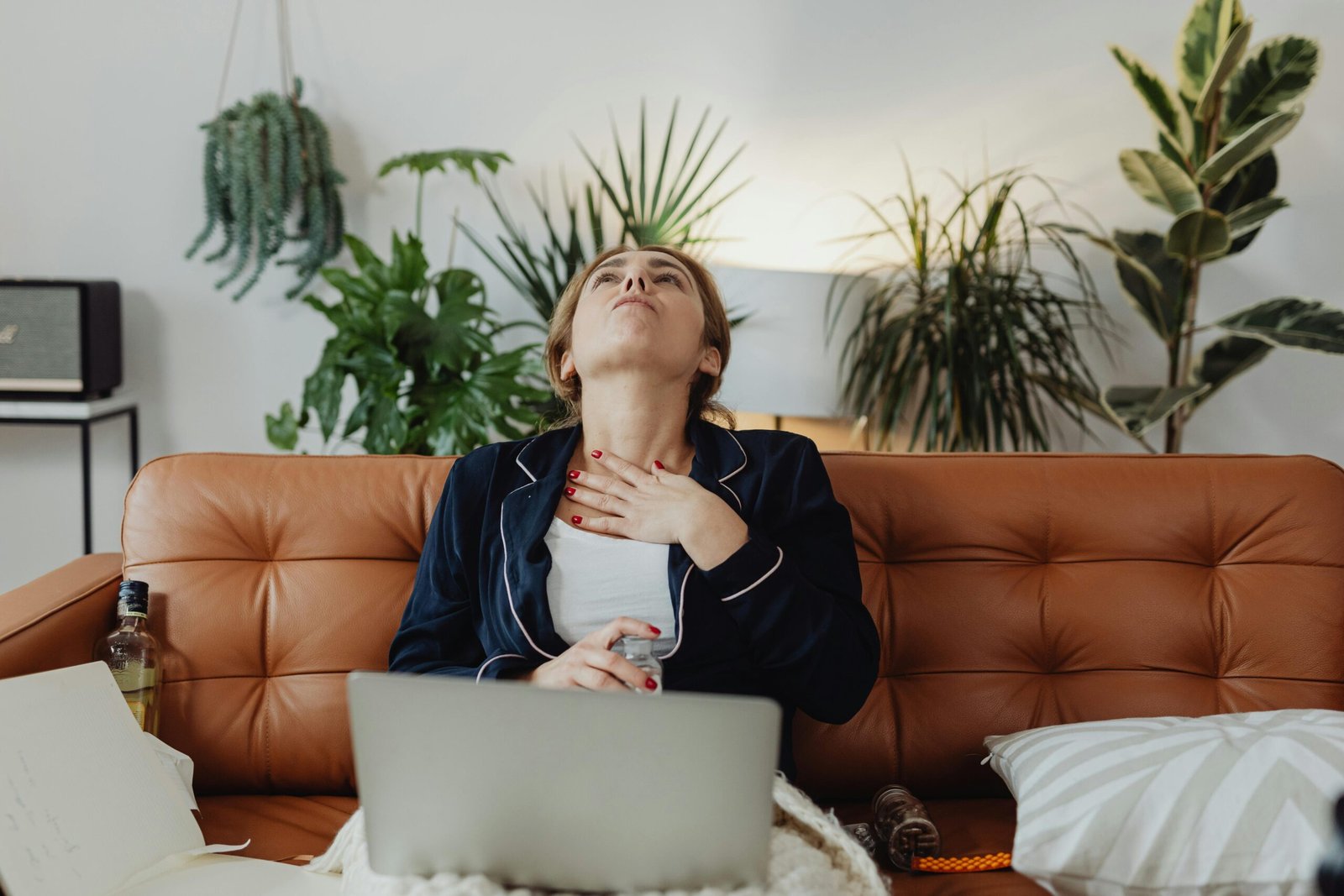 A woman sits on a brown leather couch, wearing a dark blue pajama set with white trim. She tilts her head back and places her hand on her chest while holding a small bottle, possibly medication or an inhaler. Her facial expression suggests discomfort or distress. In front of her is an open laptop, and a notebook with handwritten notes is on the couch. A bottle of alcohol and some pills are also visible nearby. The background features green houseplants, a white wall, and a speaker, giving the room a cozy yet cluttered feel.