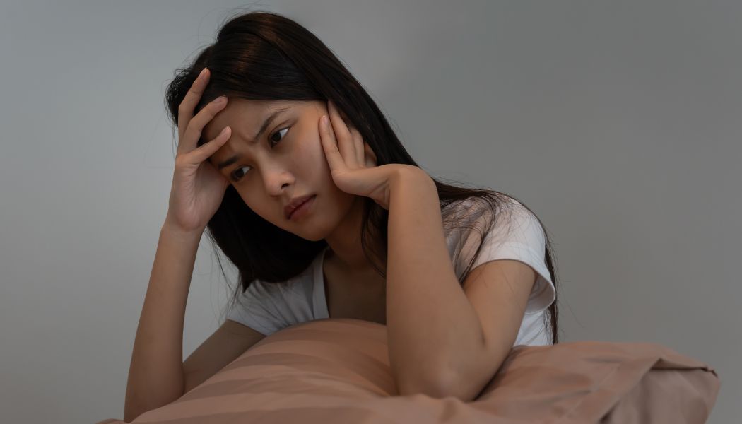 Woman sitting on a bed with her head resting on her hand appearing deep in thought.