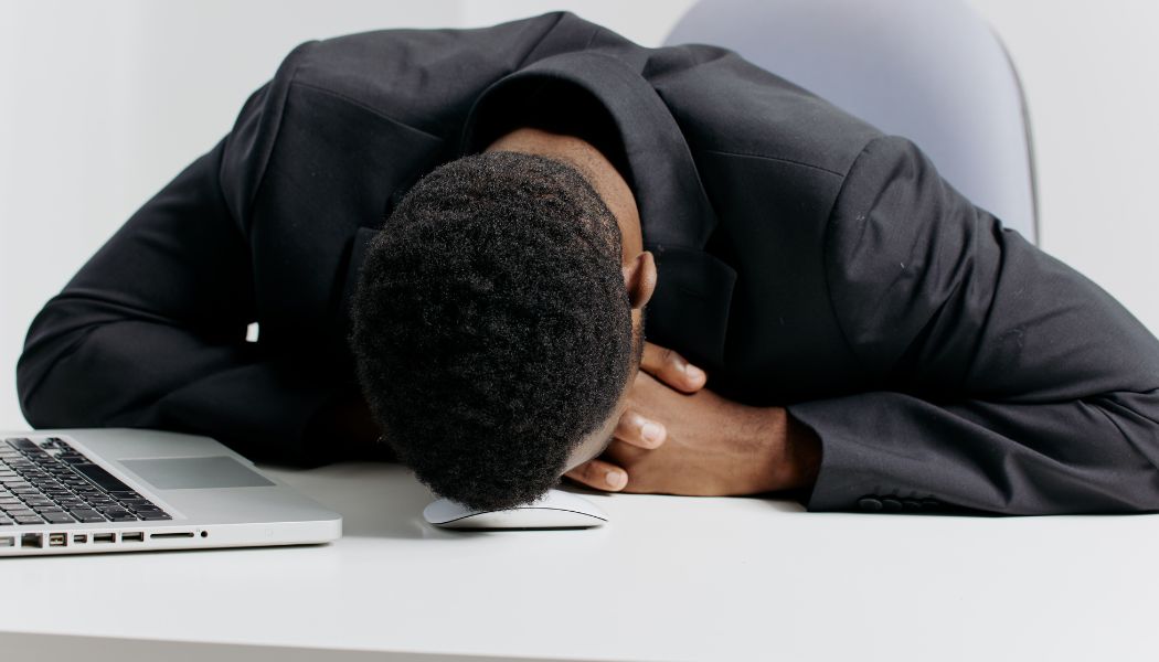 Man resting his head on a desk beside a laptop, appearing mentally exhausted