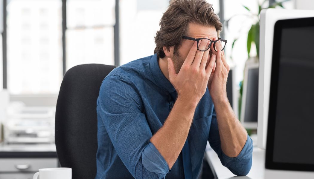 Man in glasses sitting at his desk rubbing his eyes from stress
