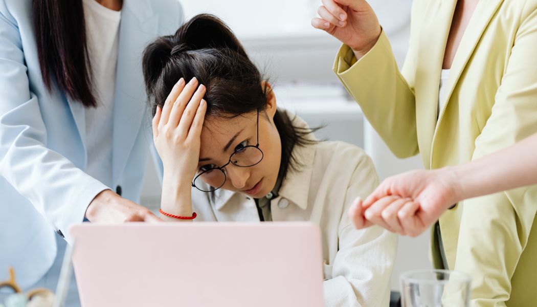 Woman sitting at a desk holding her head while coworkers gesture and point around her.