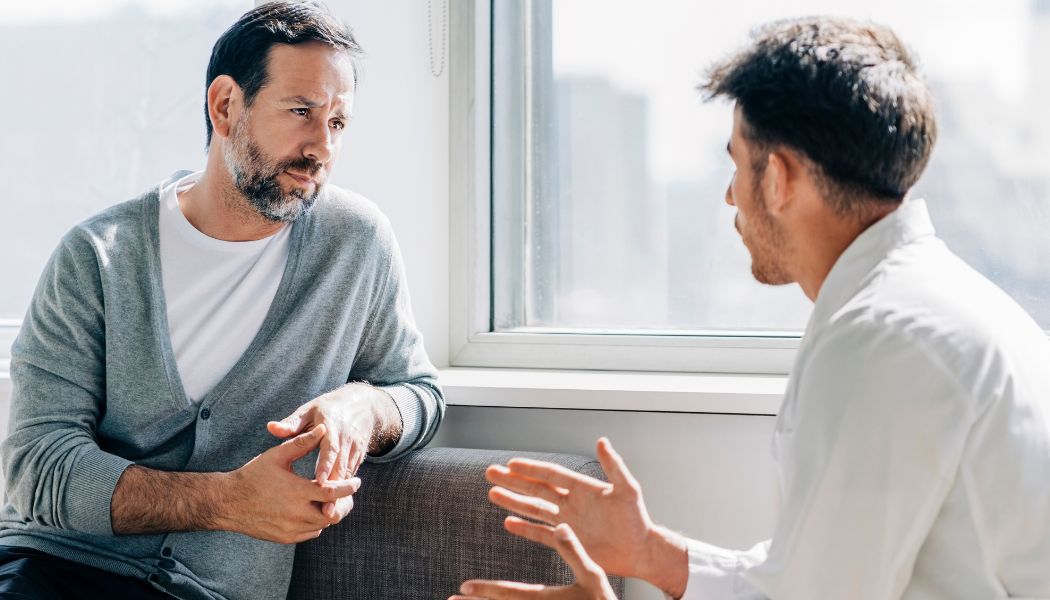 Two men sitting by a window talking seriously during a therapy session.
