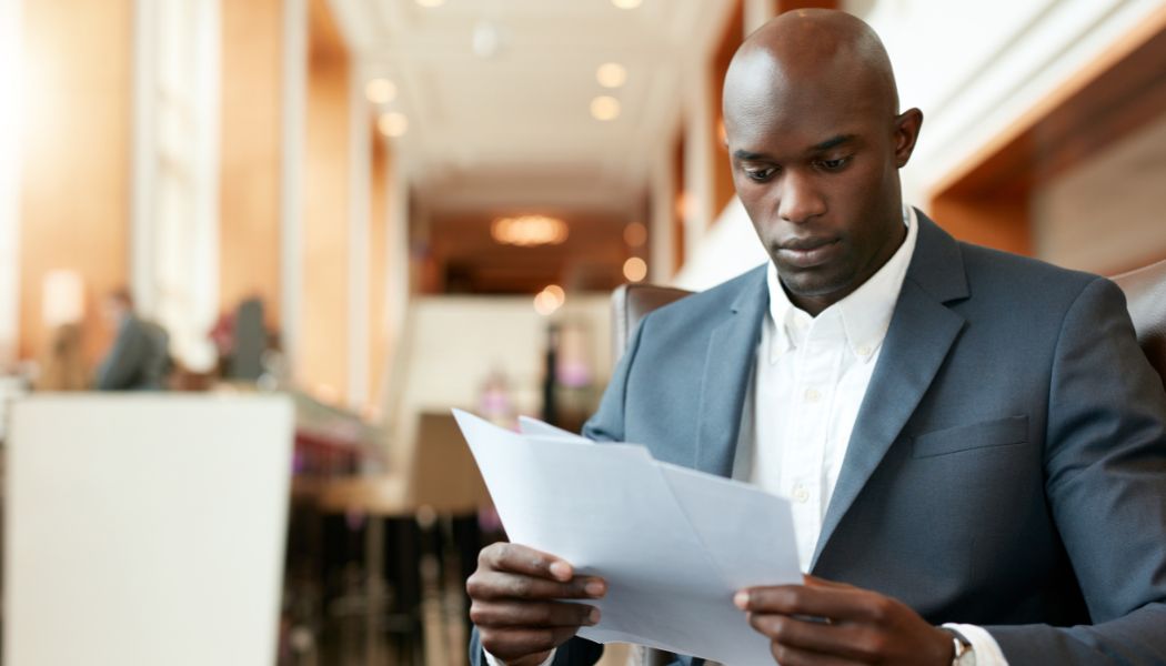 Man in a suit reading papers with focused attention in a bright lobby.