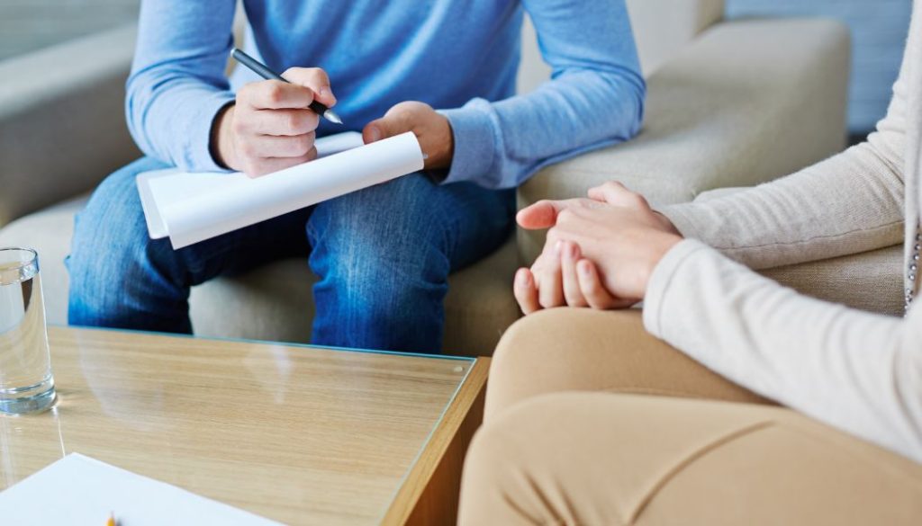 Therapist taking notes while a patient sits with clasped hands during session.